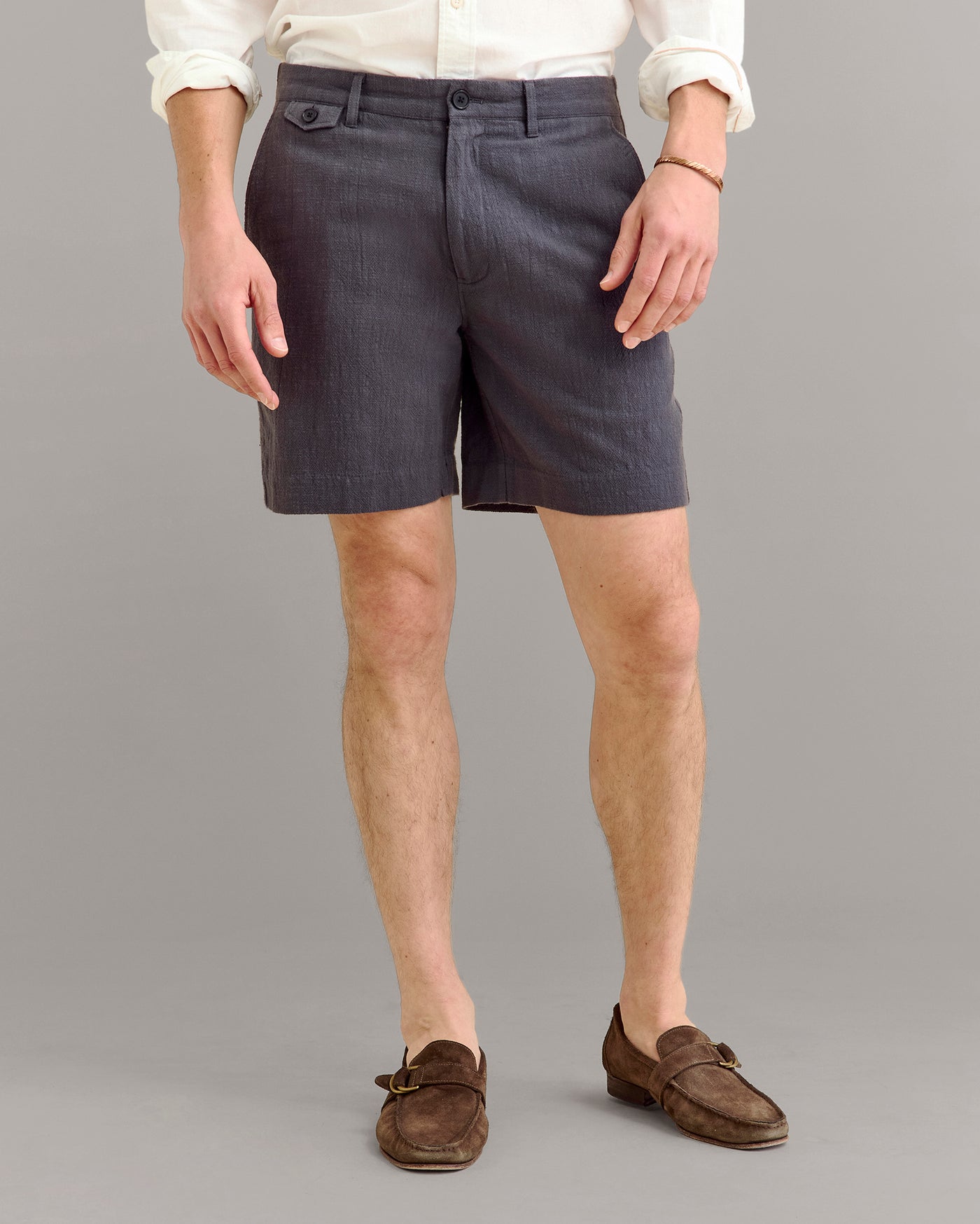 A person wearing a white long-sleeve shirt, dark gray tailored shorts, and brown loafers stands against a plain gray background. The persons face is not visible.