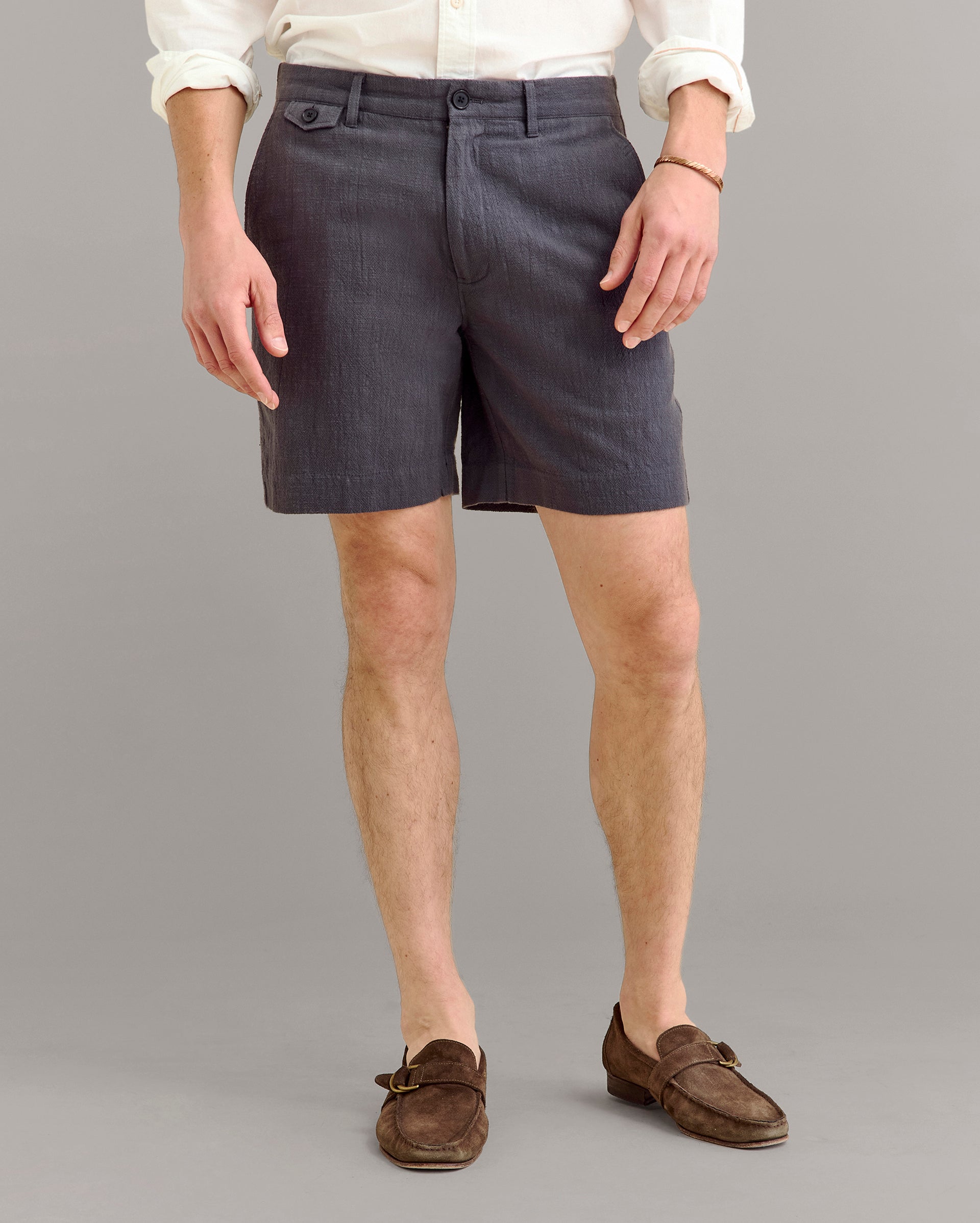 A person wearing a white long-sleeve shirt, dark gray tailored shorts, and brown loafers stands against a plain gray background. The persons face is not visible.