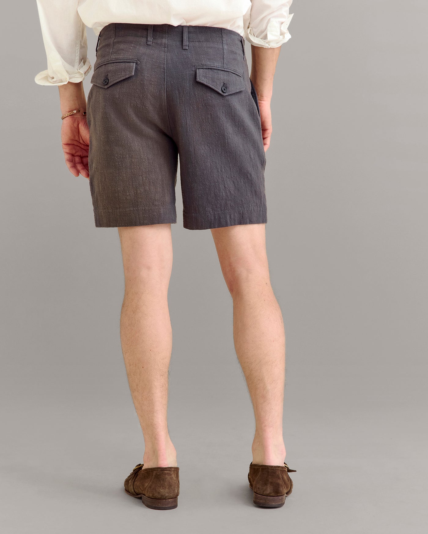 A person wearing a white long-sleeve shirt, gray tailored shorts with buttoned back pockets, and brown suede loafers, standing against a plain gray background, shown from the back.