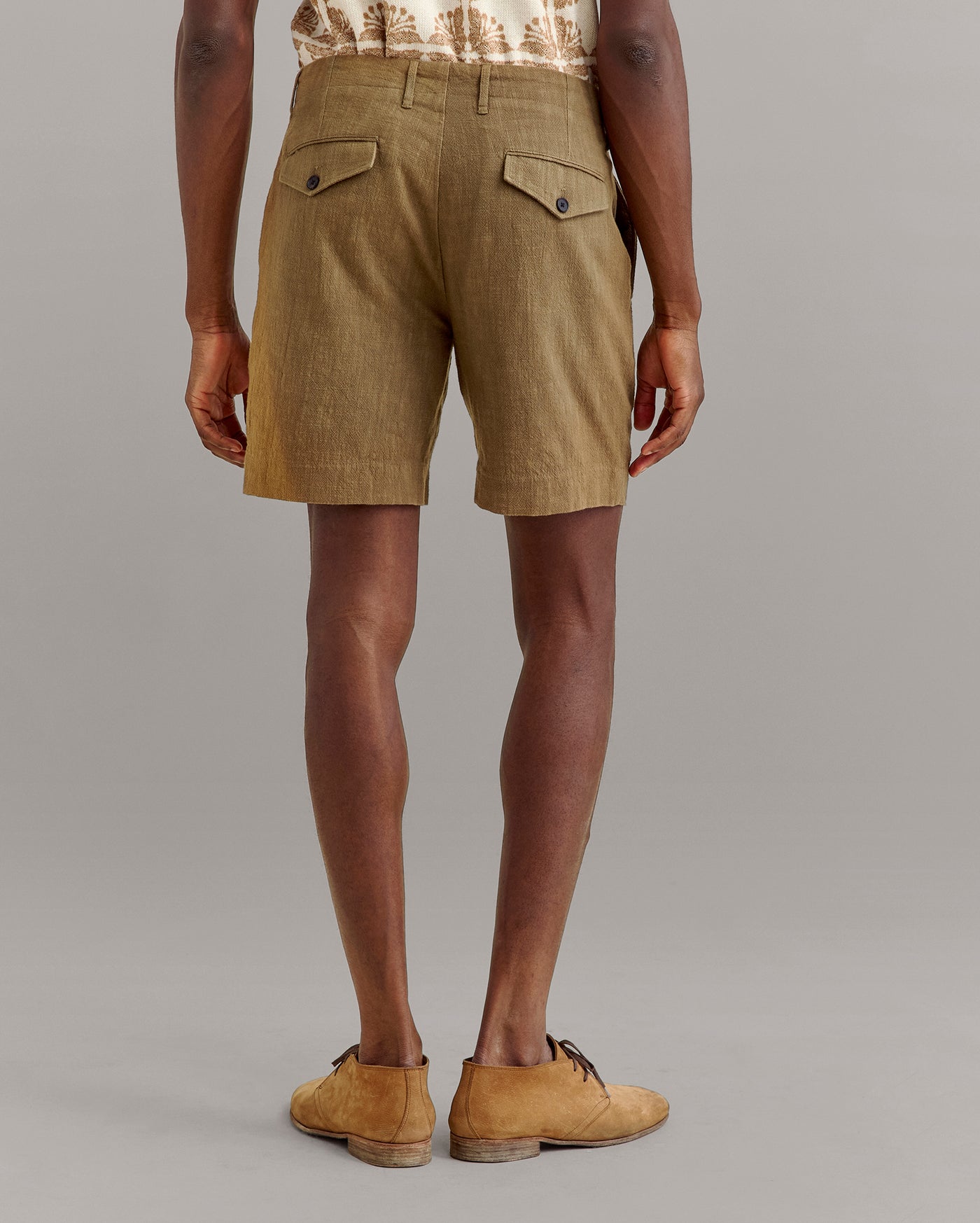 A person stands facing away, wearing light brown shorts with buttoned back pockets, a tan patterned shirt, and tan lace-up shoes against a plain gray background.