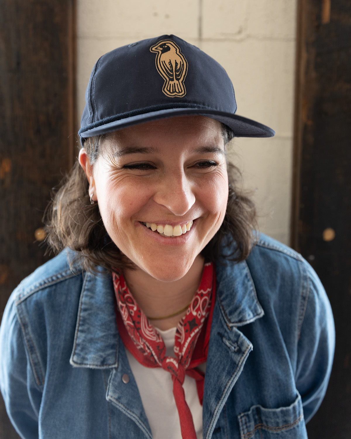 A person wearing a navy cap with a bird logo, a denim jacket, a red bandana, and a white shirt smiles while looking slightly to the side in a casual indoor setting.