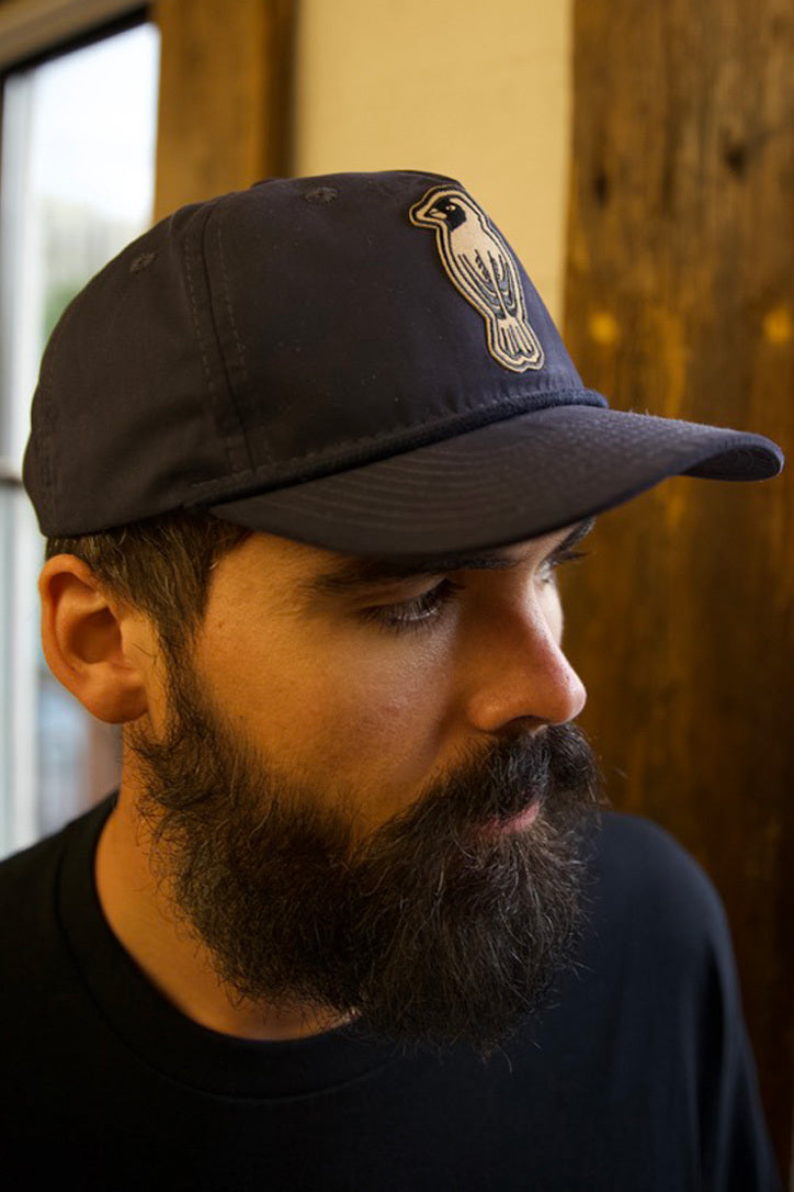 A bearded man wearing a dark baseball cap with a bird emblem looks downward. He is indoors, standing near a wooden beam and a window, dressed in a black shirt.