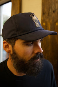 A bearded man wearing a dark baseball cap with a bird emblem looks downward. He is indoors, standing near a wooden beam and a window, dressed in a black shirt.