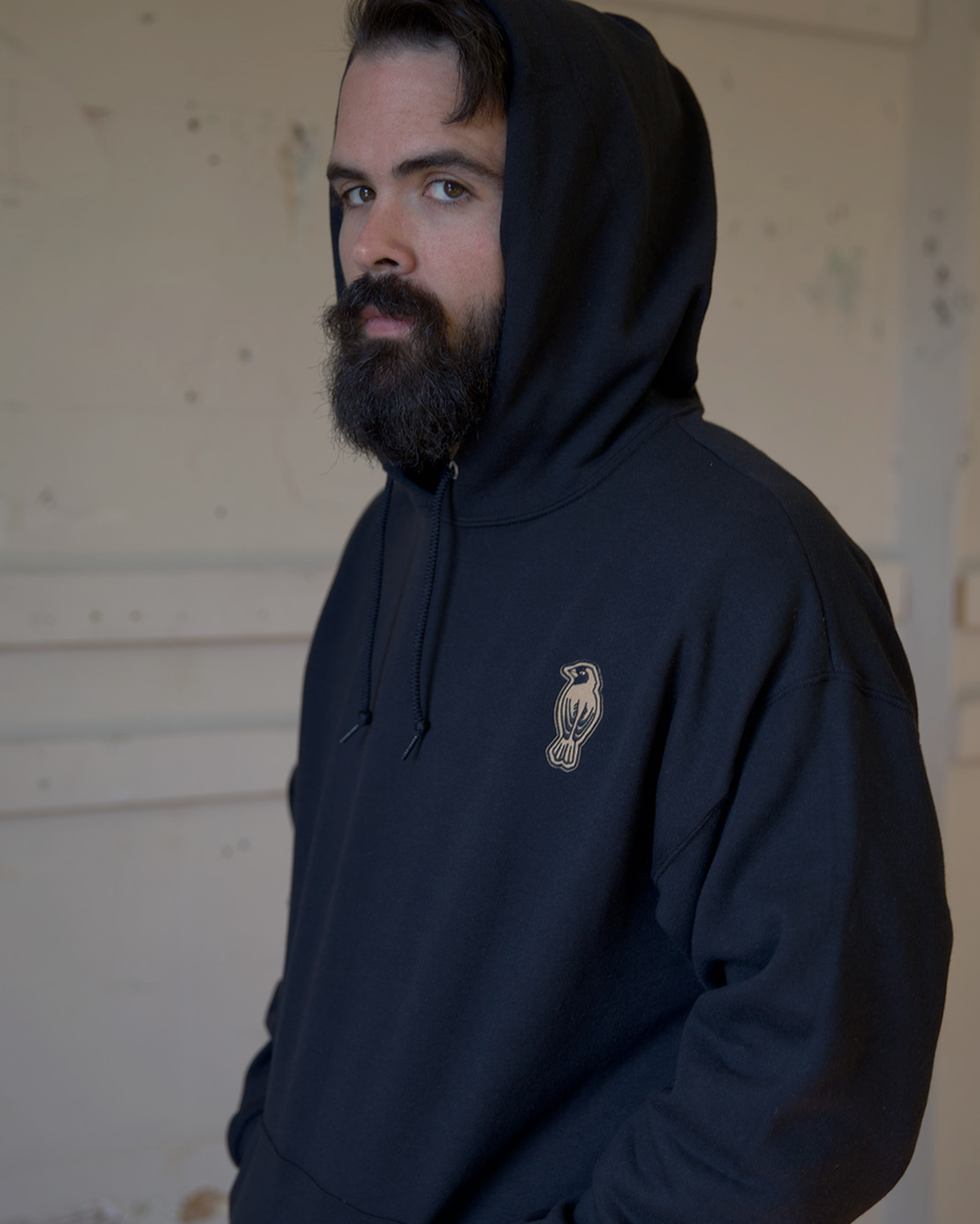 A man with a dark beard and mustache wears a black hooded sweatshirt with a small owl logo on the chest, standing against a light-colored, textured wall.