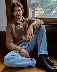 A man with medium-length brown hair and a beard sits on a wooden floor by a large window, wearing a tan shirt, light blue jeans, and black boots, looking calmly at the camera.