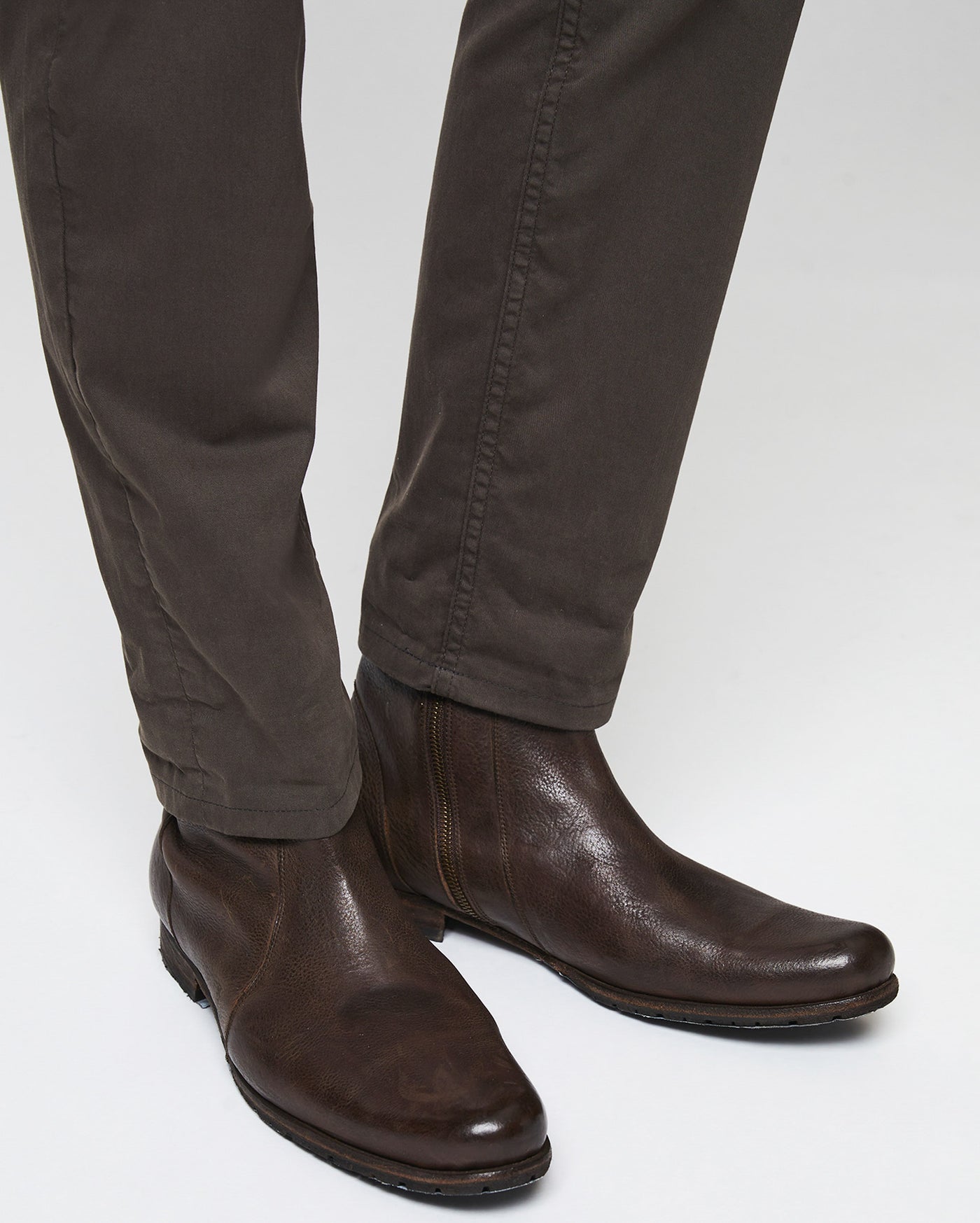 A person wearing dark brown leather ankle boots and dark brown pants, shown from the knees down against a plain, light-colored background.