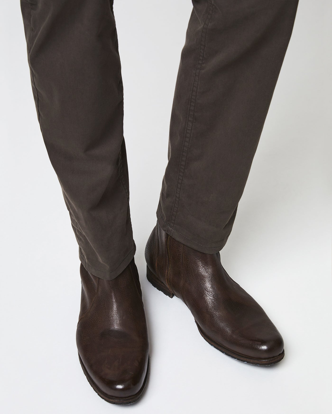 A person wearing dark brown leather Chelsea boots and dark brown pants standing on a light-colored surface.