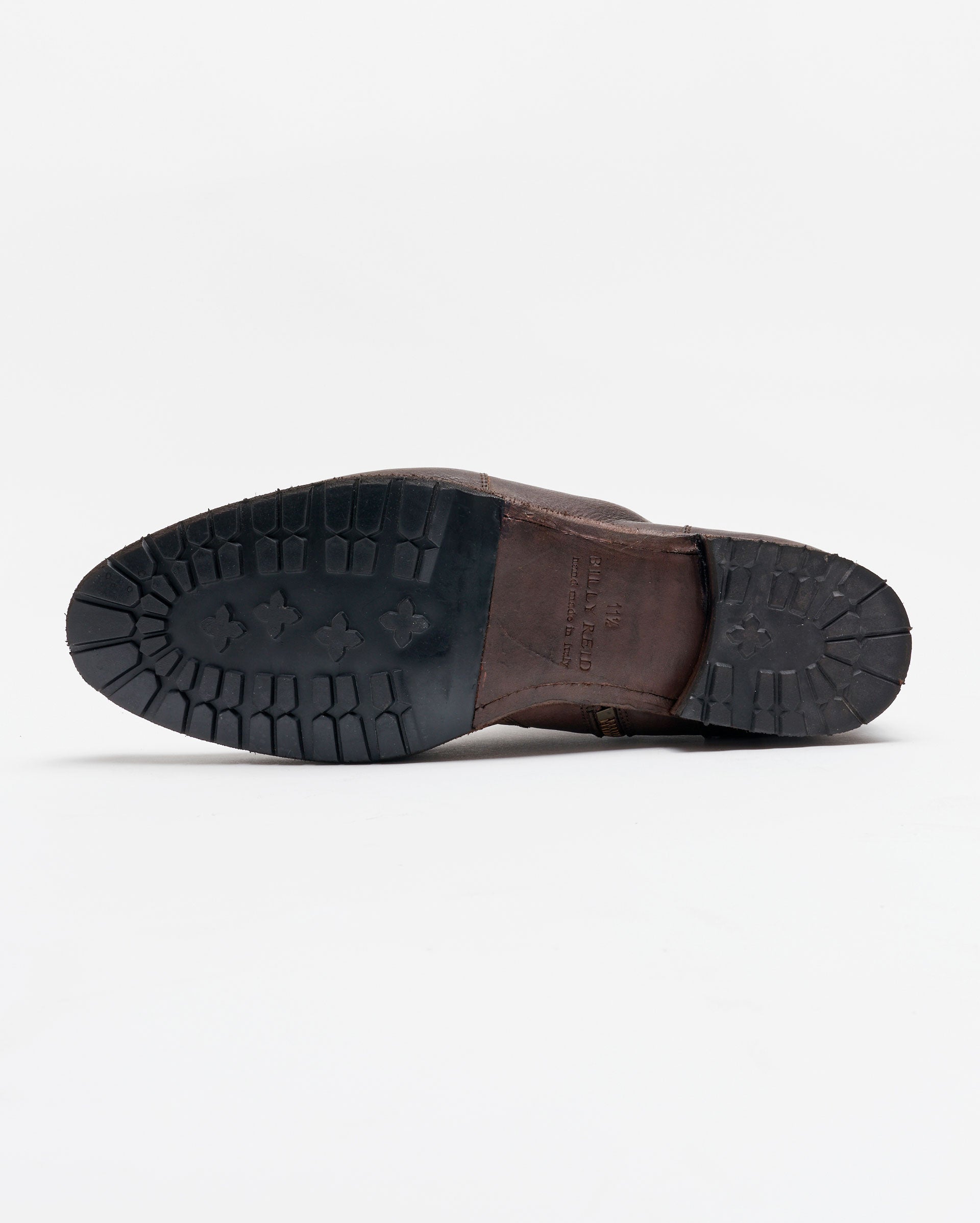 A brown leather shoe lying on its side, showing the black rubber sole with a rugged tread pattern, against a plain white background.
