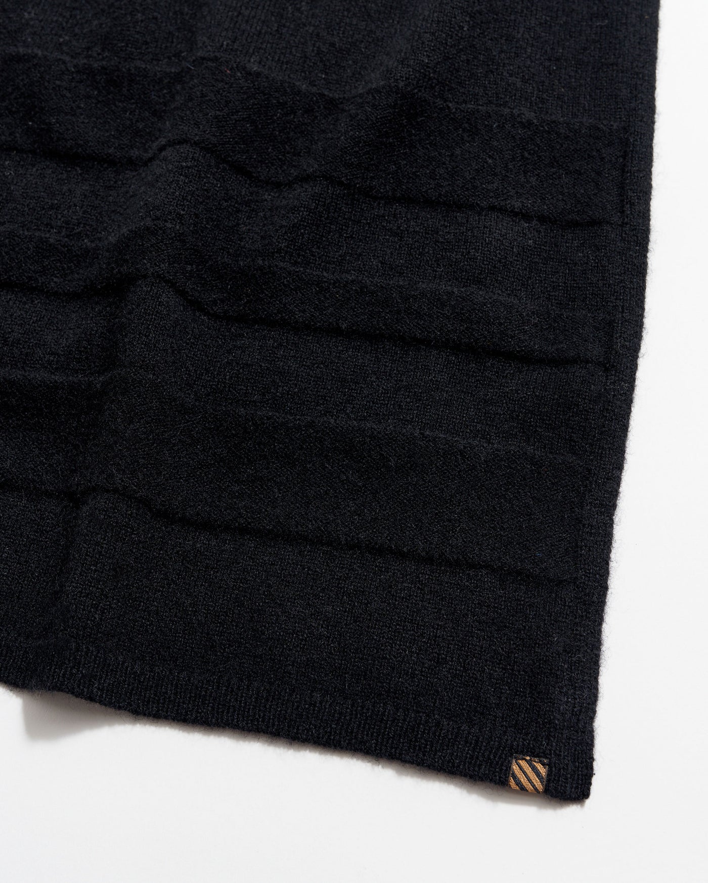 A close-up of a black knitted fabric with horizontal textured stripes and a small brown tag at the bottom corner, photographed on a white background.