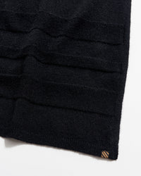 A close-up of a black knitted fabric with horizontal textured stripes and a small brown tag at the bottom corner, photographed on a white background.