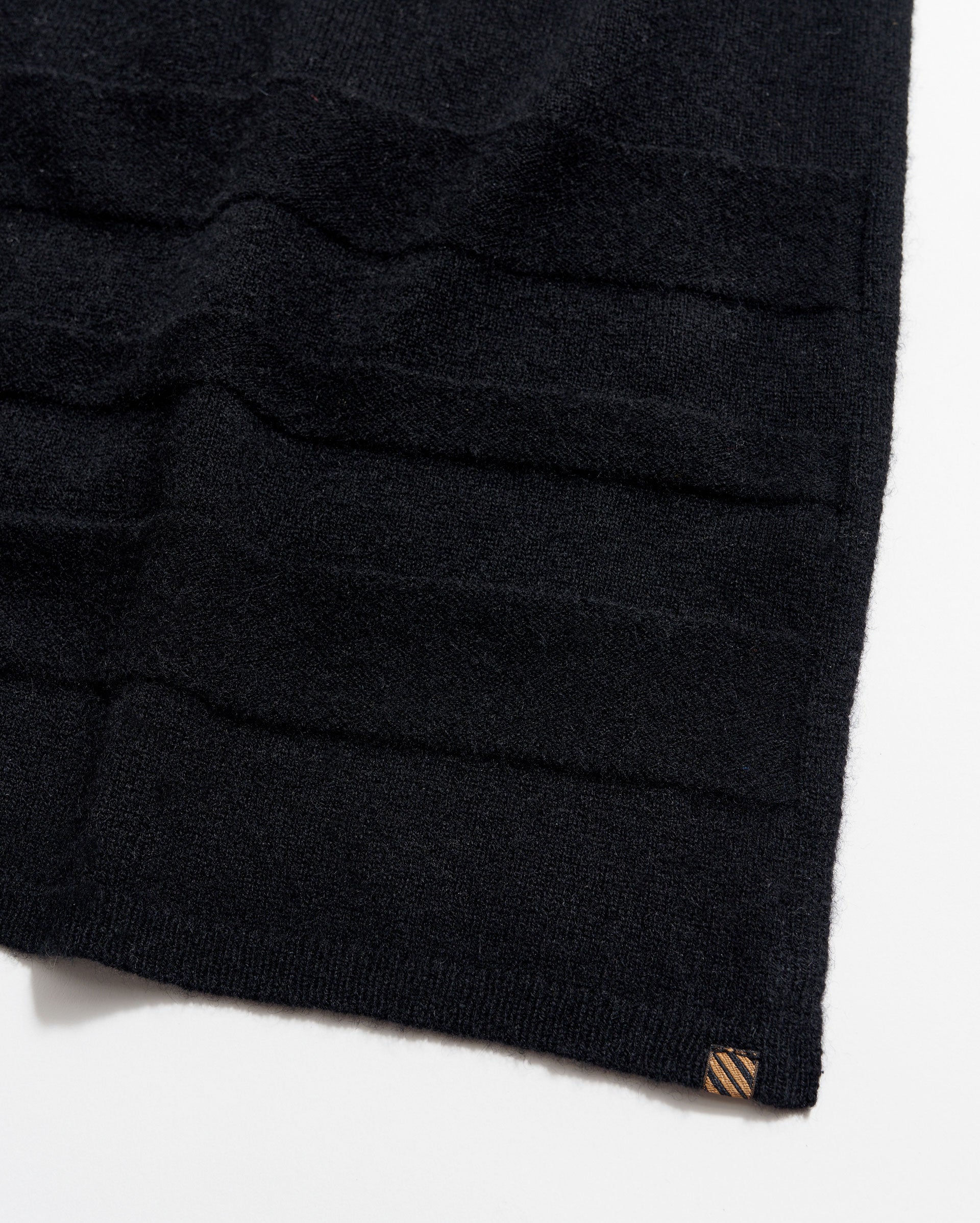 A close-up of a black knitted fabric with horizontal textured stripes and a small brown tag at the bottom corner, photographed on a white background.