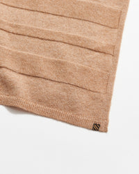 A close-up of a light brown knitted fabric with horizontal textured stripes, finished with a ribbed edge and a small rectangular tag in the corner on a white background.