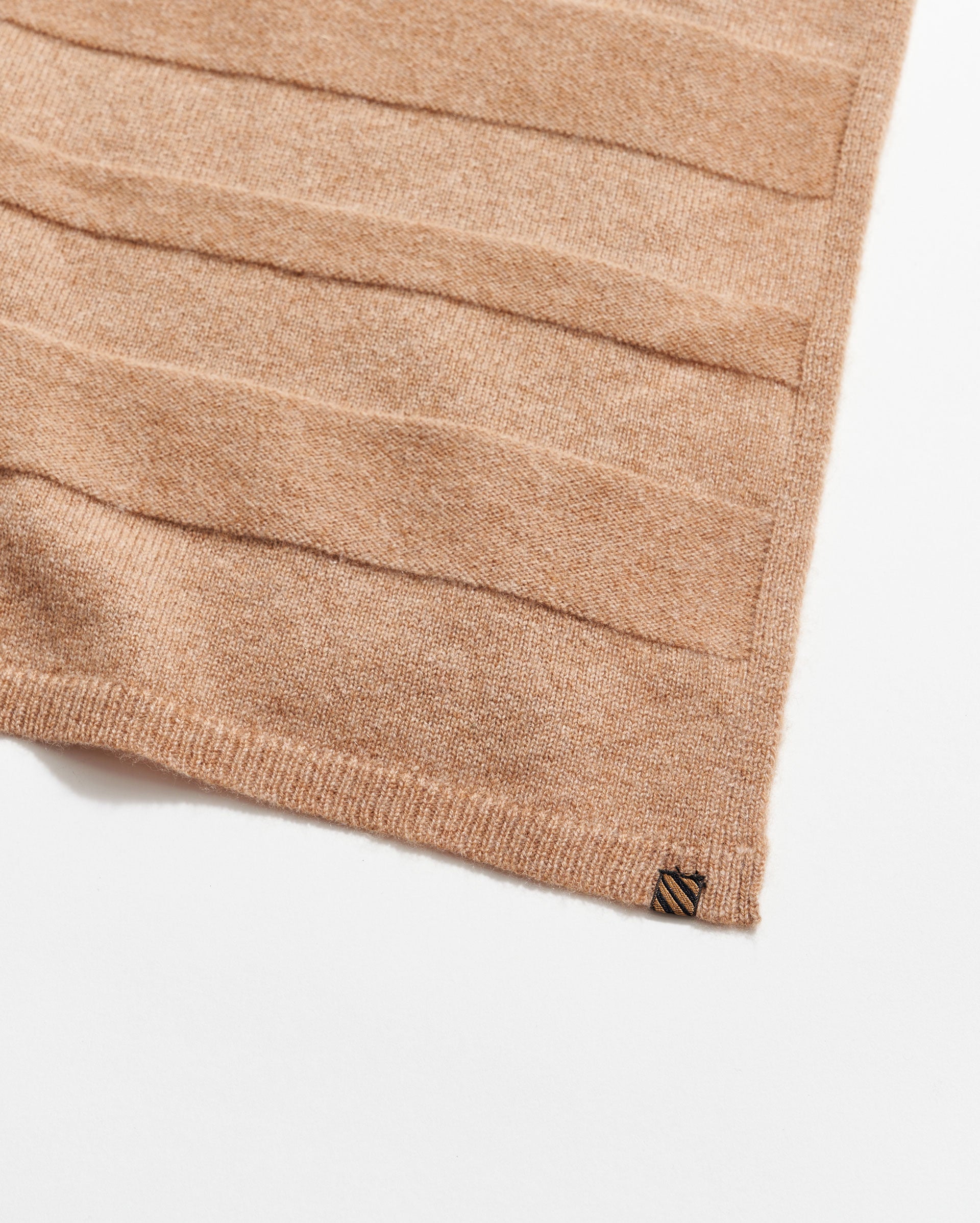 A close-up of a light brown knitted fabric with horizontal textured stripes, finished with a ribbed edge and a small rectangular tag in the corner on a white background.