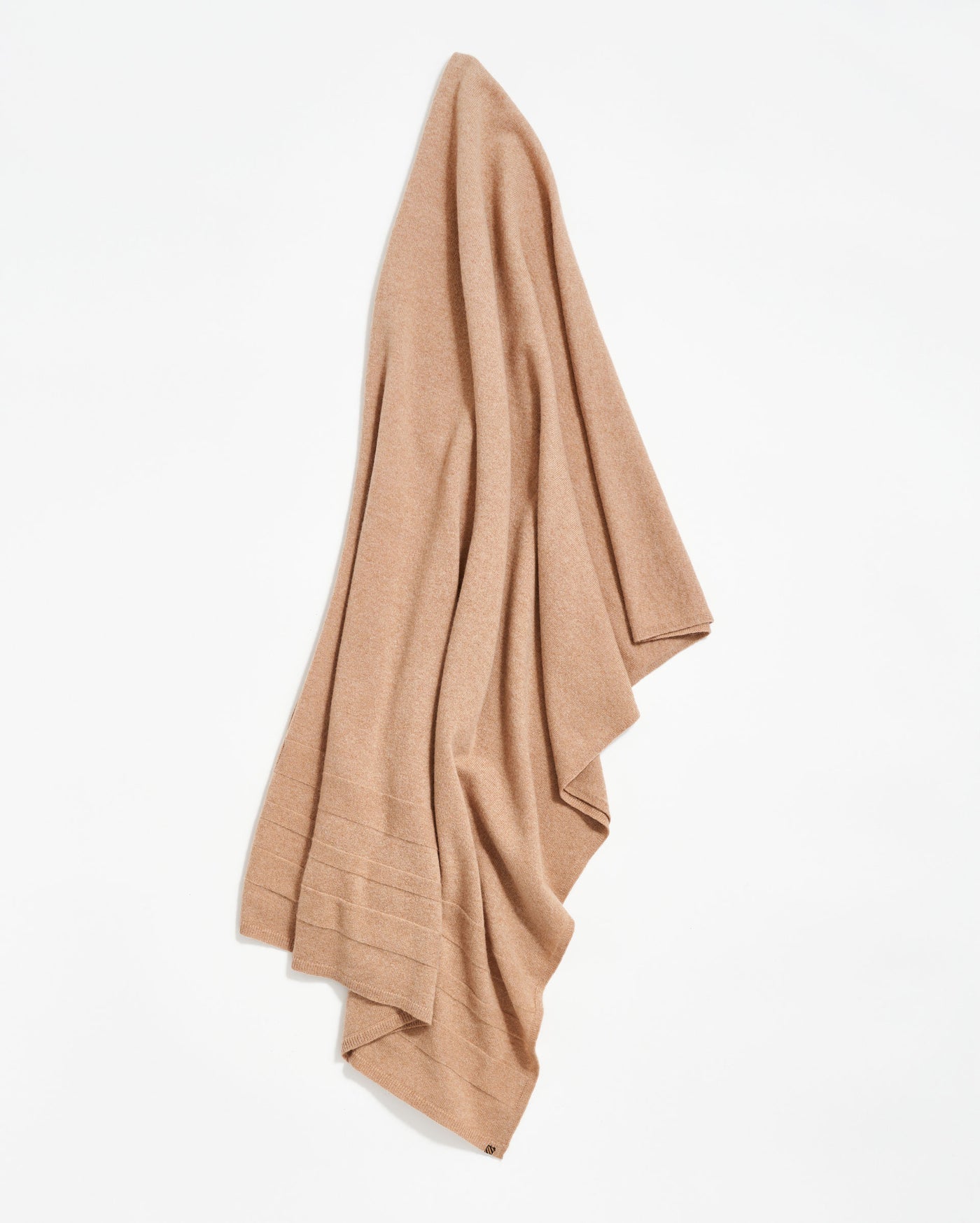 A beige or light brown blanket or throw is draped against a white background, displaying soft, slightly textured fabric with subtle horizontal stitched lines near the bottom edge.