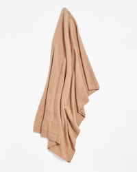 A beige or light brown blanket or throw is draped against a white background, displaying soft, slightly textured fabric with subtle horizontal stitched lines near the bottom edge.
