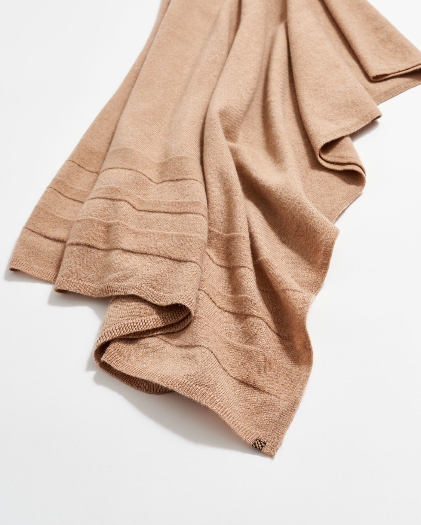 A soft, light brown cashmere scarf with subtle horizontal ribbed stripes, laid out on a white surface. A small metallic tag is attached to one corner.