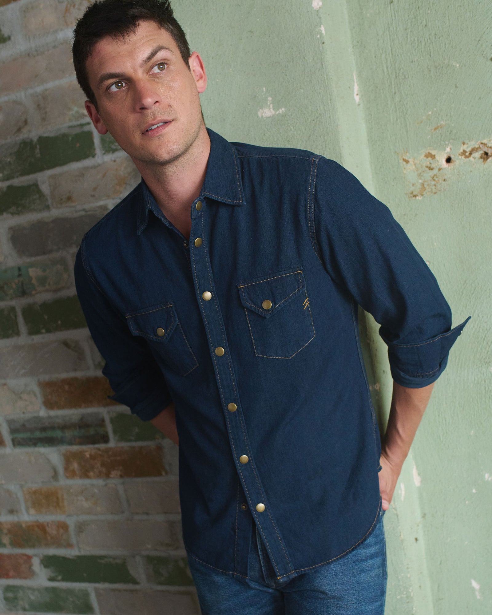 A man in a dark denim shirt and jeans stands against a green, weathered wall with exposed brick, looking slightly upward with a thoughtful expression.