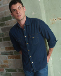 A man in a dark denim shirt and jeans stands against a green, weathered wall with exposed brick, looking slightly upward with a thoughtful expression.