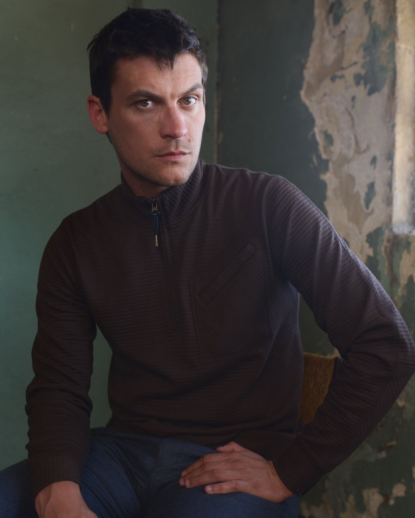 A man with short dark hair, wearing a dark brown zip-up sweater and blue jeans, sits on a chair against a textured wall with peeling paint, looking directly at the camera with a neutral expression.