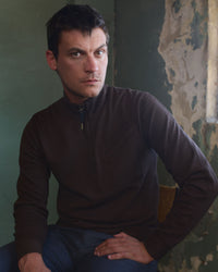 A man with short dark hair, wearing a dark brown zip-up sweater and blue jeans, sits on a chair against a textured wall with peeling paint, looking directly at the camera with a neutral expression.