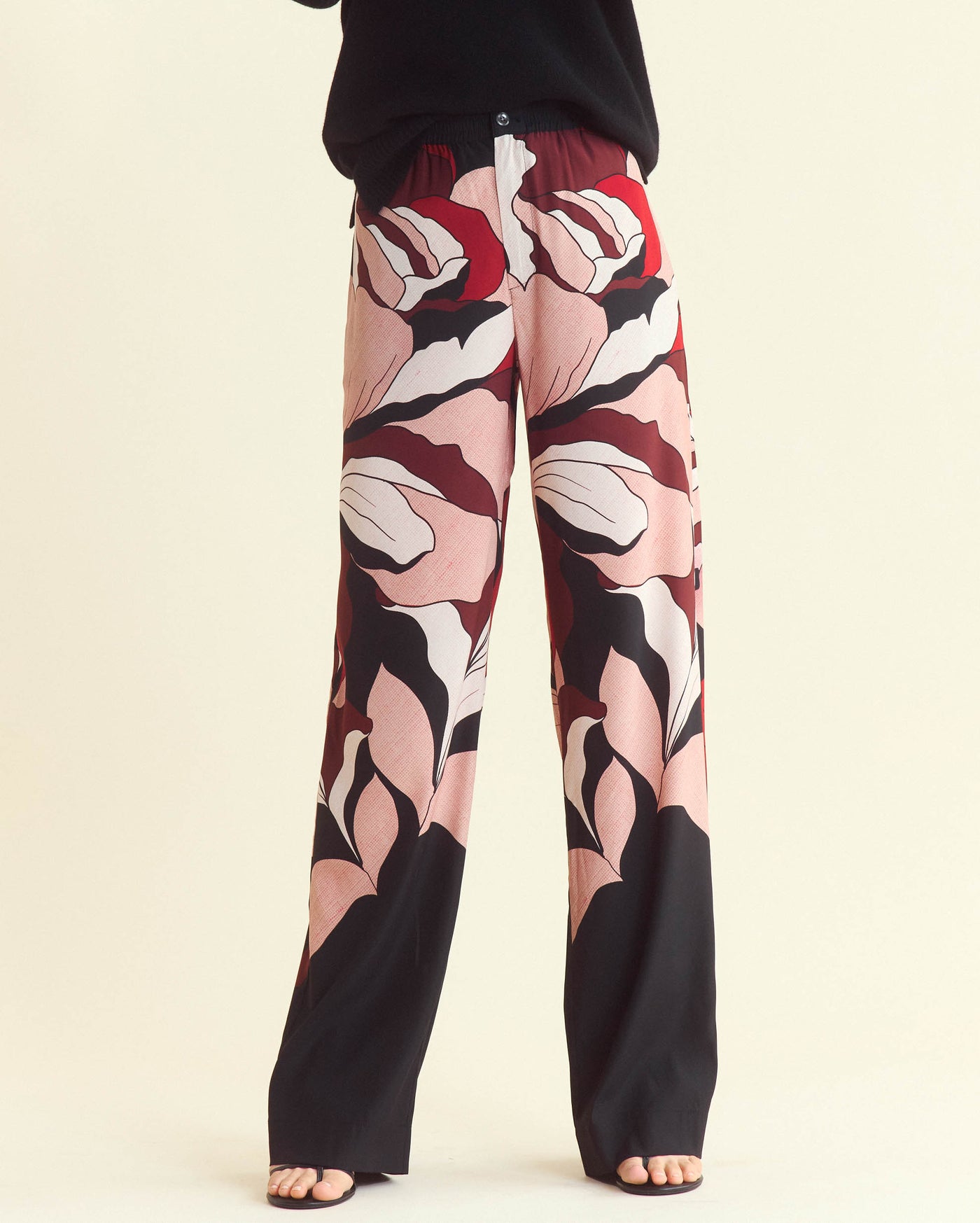 Person wearing wide-leg pants with a bold, abstract pink, red, black, and white floral pattern. Only the lower half of the body is visible; feet are in sandals and the background is light beige.