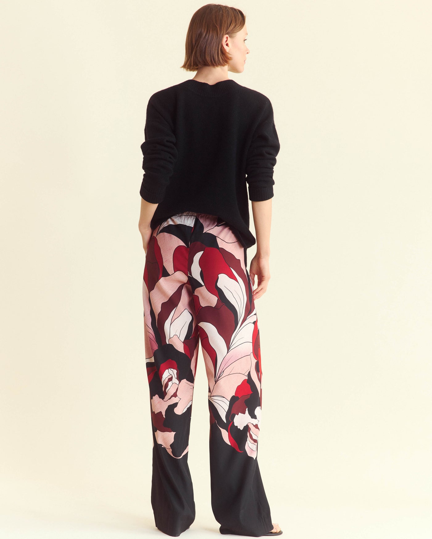 A person stands facing away, wearing a black long-sleeve top and loose, wide-leg pants with a bold, abstract floral pattern in red, pink, white, and black. The background is a plain off-white color.