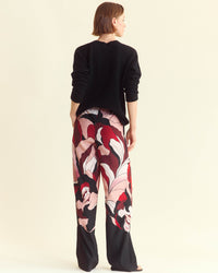 A person stands facing away, wearing a black long-sleeve top and loose, wide-leg pants with a bold, abstract floral pattern in red, pink, white, and black. The background is a plain off-white color.