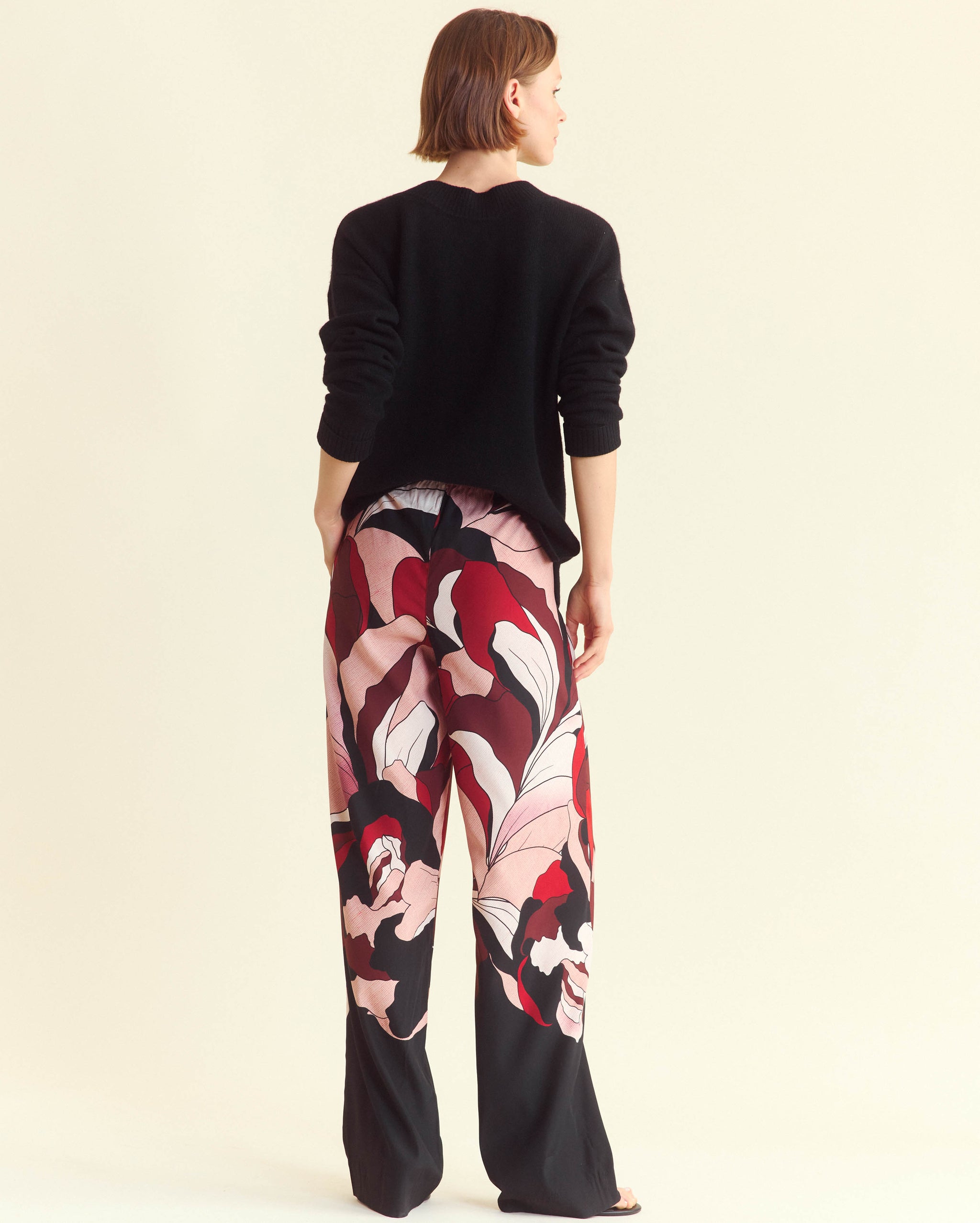 A person stands facing away, wearing a black long-sleeve top and loose, wide-leg pants with a bold, abstract floral pattern in red, pink, white, and black. The background is a plain off-white color.