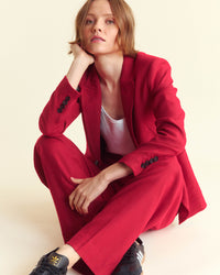 A person with short light brown hair sits on the floor wearing a bright red suit, white shirt, and black sneakers. They rest their head on one hand and look directly at the camera with a neutral expression.