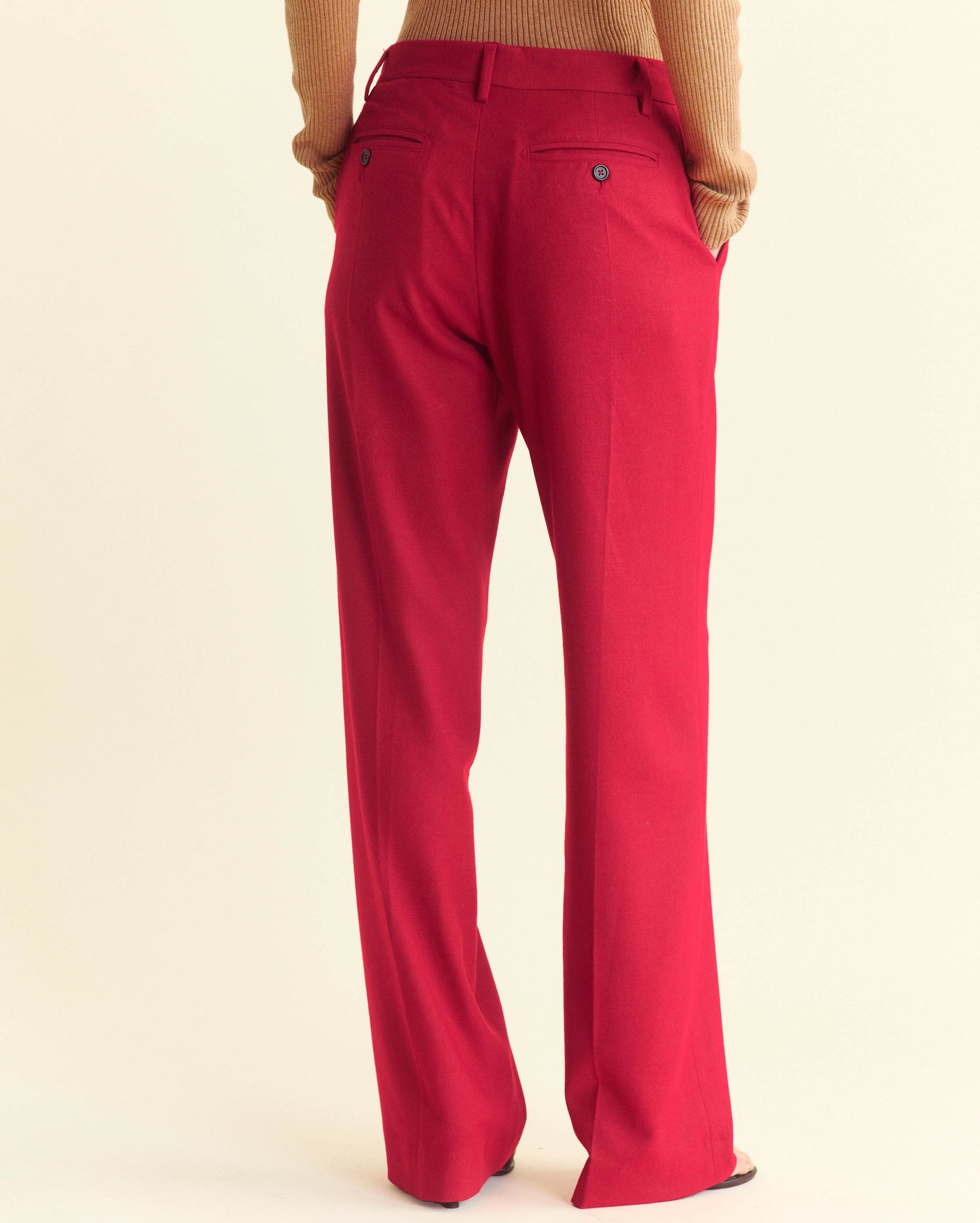 A person wearing bright red dress pants and a light brown ribbed sweater is standing with their back to the camera against a plain light background.