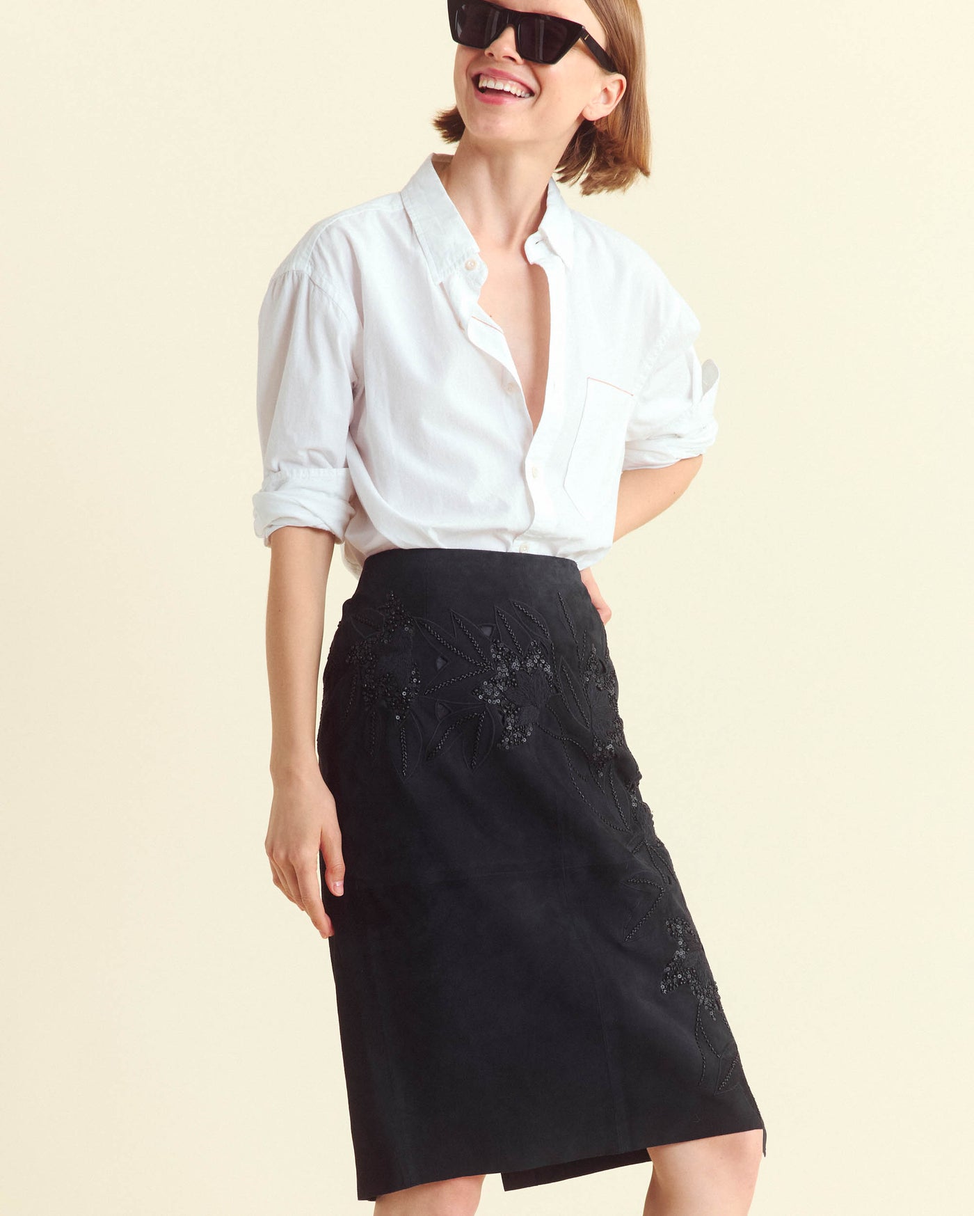A person wearing a white button-up shirt, black skirt with subtle embroidery, and large black sunglasses smiles while looking to the side against a plain light background.
