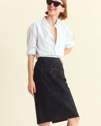 A person wearing a white button-up shirt, black skirt with subtle embroidery, and large black sunglasses smiles while looking to the side against a plain light background.