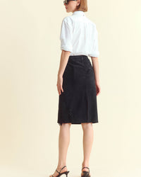 A woman stands facing away, wearing a white long-sleeve shirt with rolled-up sleeves, a black knee-length skirt with subtle embellishments, black heeled sandals, and sunglasses. The background is plain and light-colored.