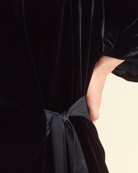 A close-up of a person wearing a black velvet garment tied at the waist with a matching black fabric sash, with one hand tucked into a pocket.