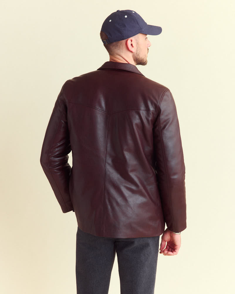 A man wearing a navy baseball cap, a dark brown leather jacket, and gray pants stands facing away from the camera against a plain light background.