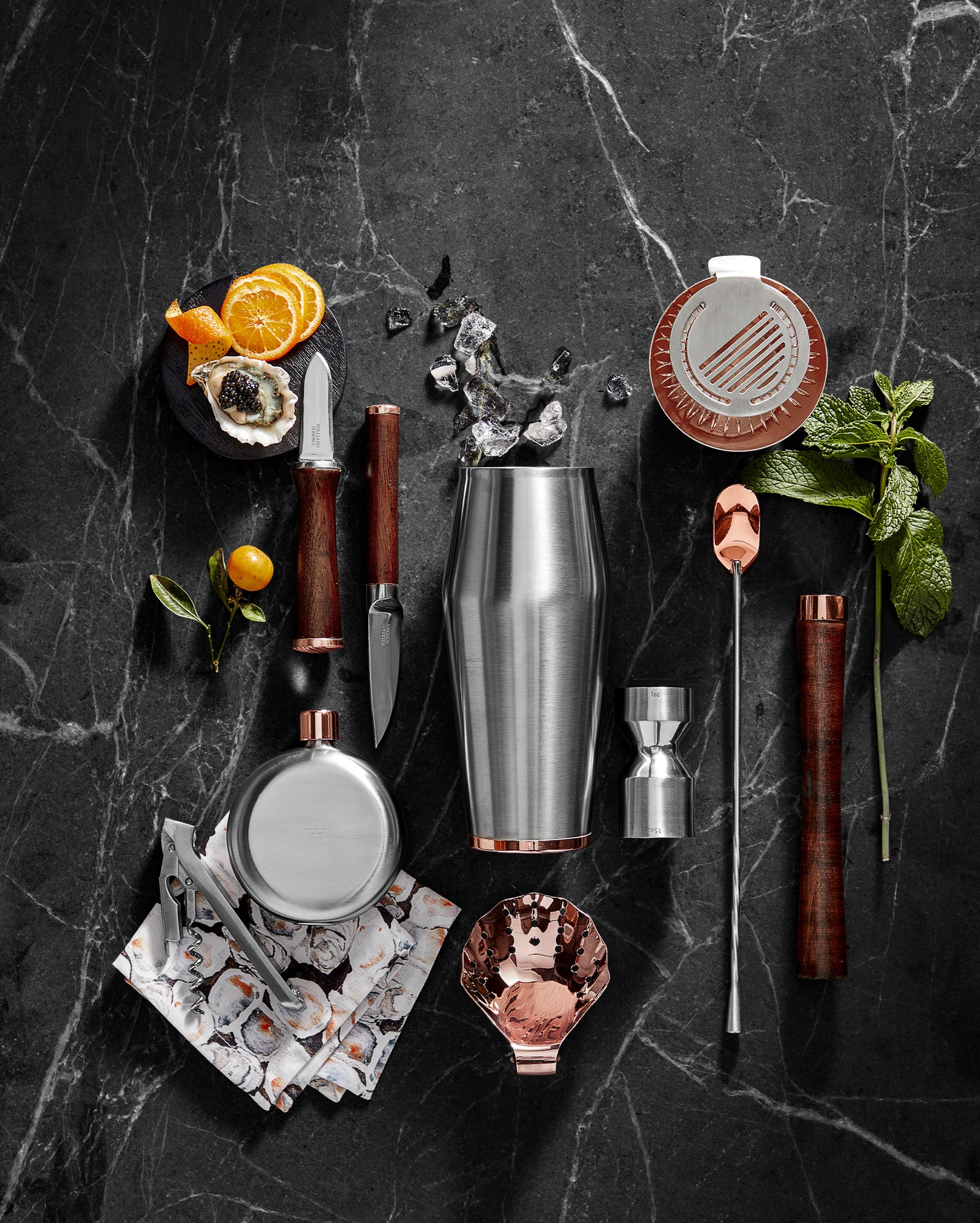 A cocktail set on a dark marble surface with a shaker, jigger, muddler, strainer, bar spoon, knife, peeler, flask, citrus, mint, ice, and a patterned napkin arranged neatly.