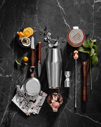 A cocktail set on a dark marble surface with a shaker, jigger, muddler, strainer, bar spoon, knife, peeler, flask, citrus, mint, ice, and a patterned napkin arranged neatly.