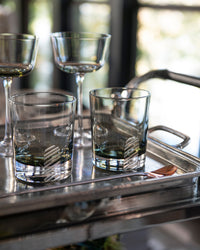 Four empty glasses—two short, two tall—sit on a metal tray with a small copper spoon. The background shows blurred windows and greenery, creating a bright and inviting atmosphere.