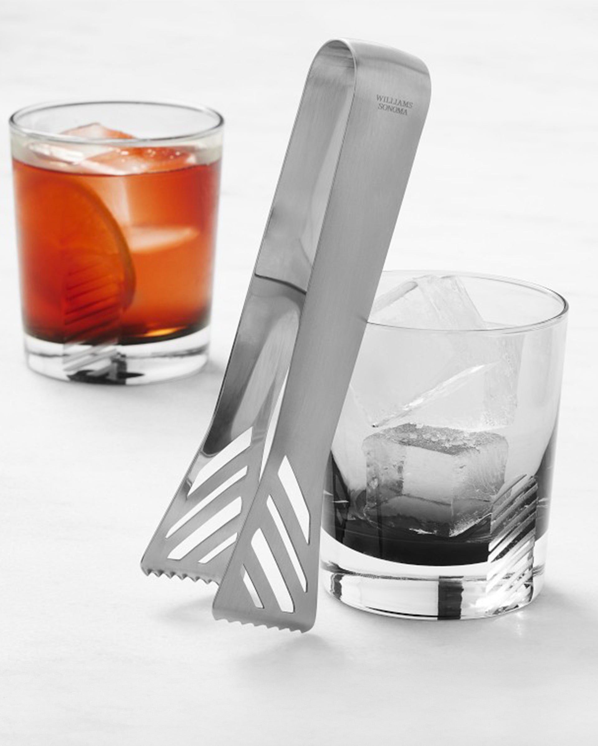 Two short glasses with large ice cubes, one containing a brown drink with an orange slice. Metal ice tongs rest on the rim of the glass with clear ice cubes on a white surface.