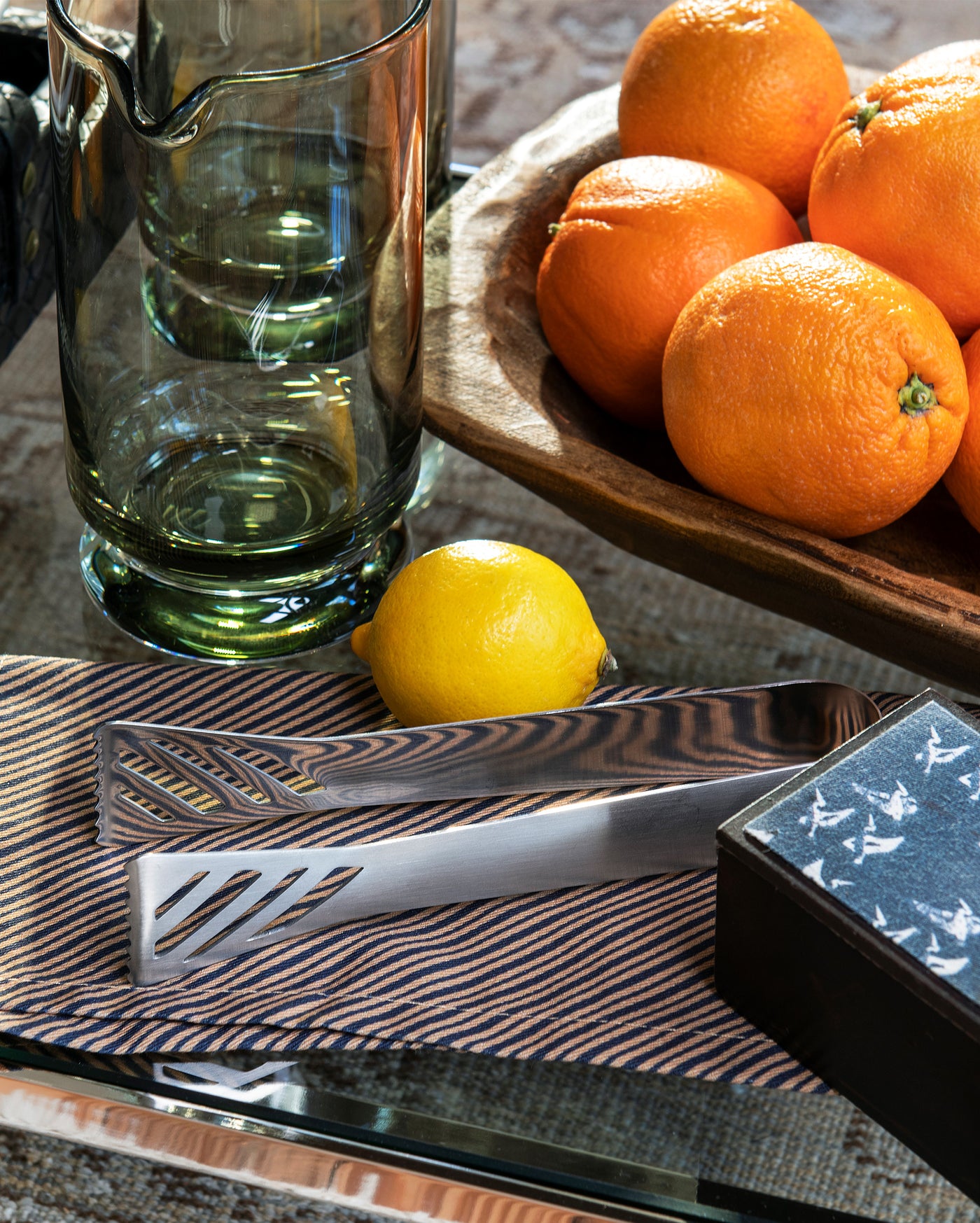 A glass pitcher, a wooden bowl of oranges, a lemon, metal utensils on a striped napkin, and a black box with a white bird pattern are arranged on a reflective surface.