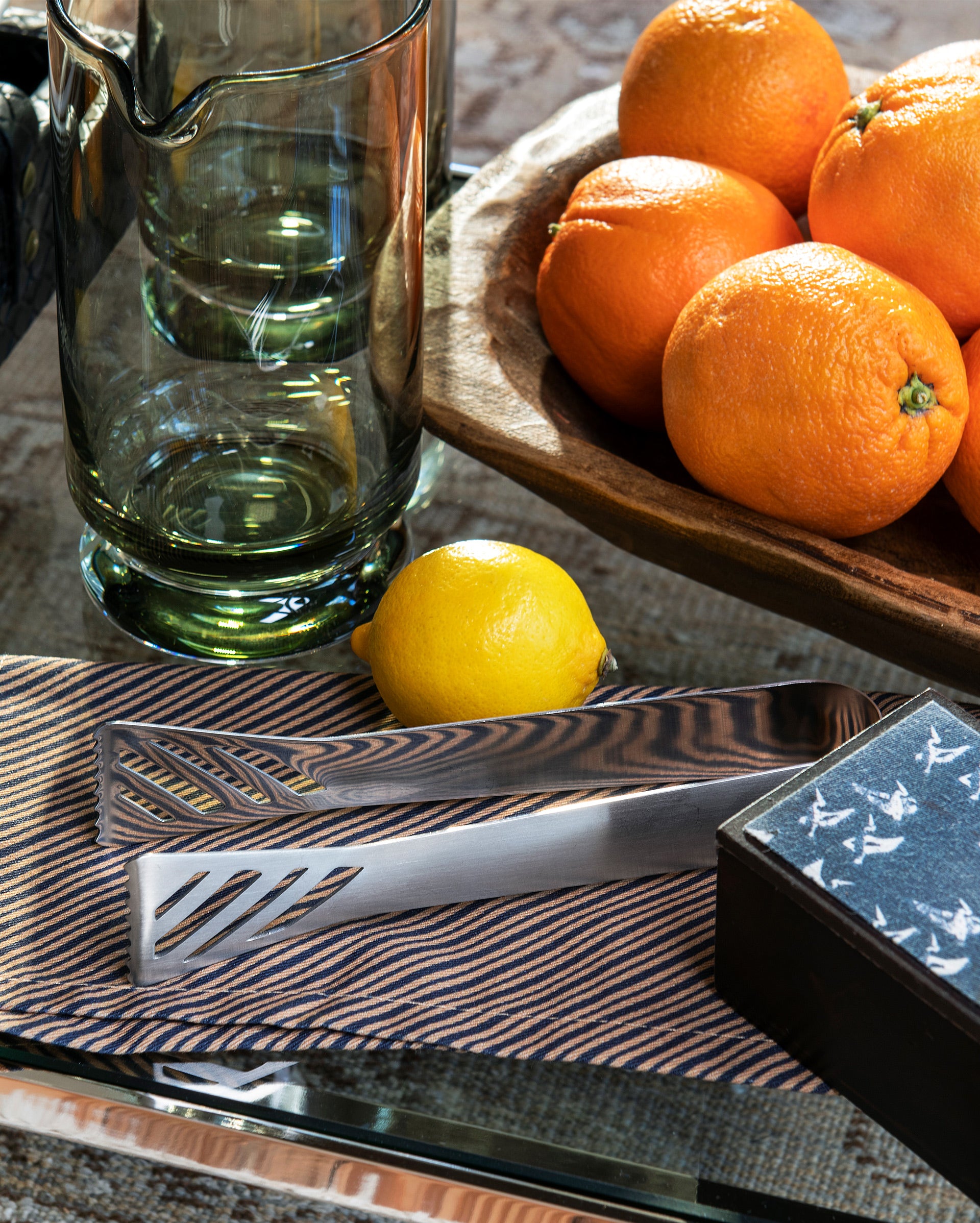 A glass pitcher, a wooden bowl of oranges, a lemon, metal utensils on a striped napkin, and a black box with a white bird pattern are arranged on a reflective surface.
