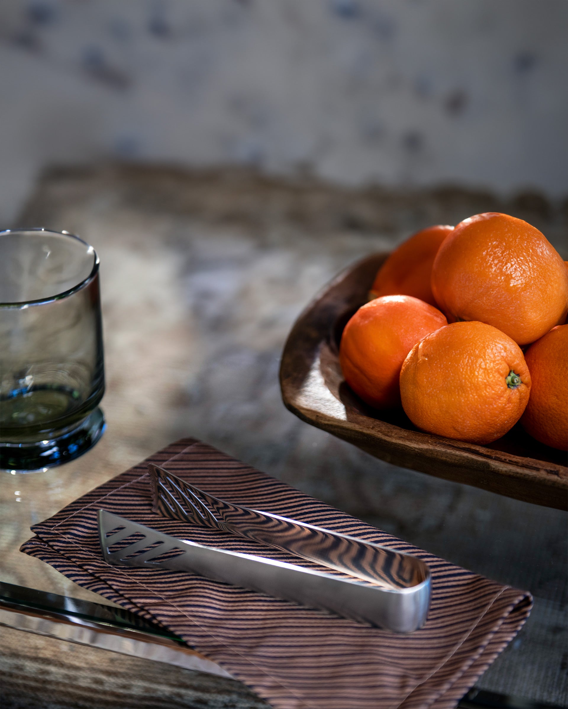 A wooden bowl filled with oranges sits on a rustic table next to a dark glass tumbler and a patterned napkin holding a silver cheese knife.