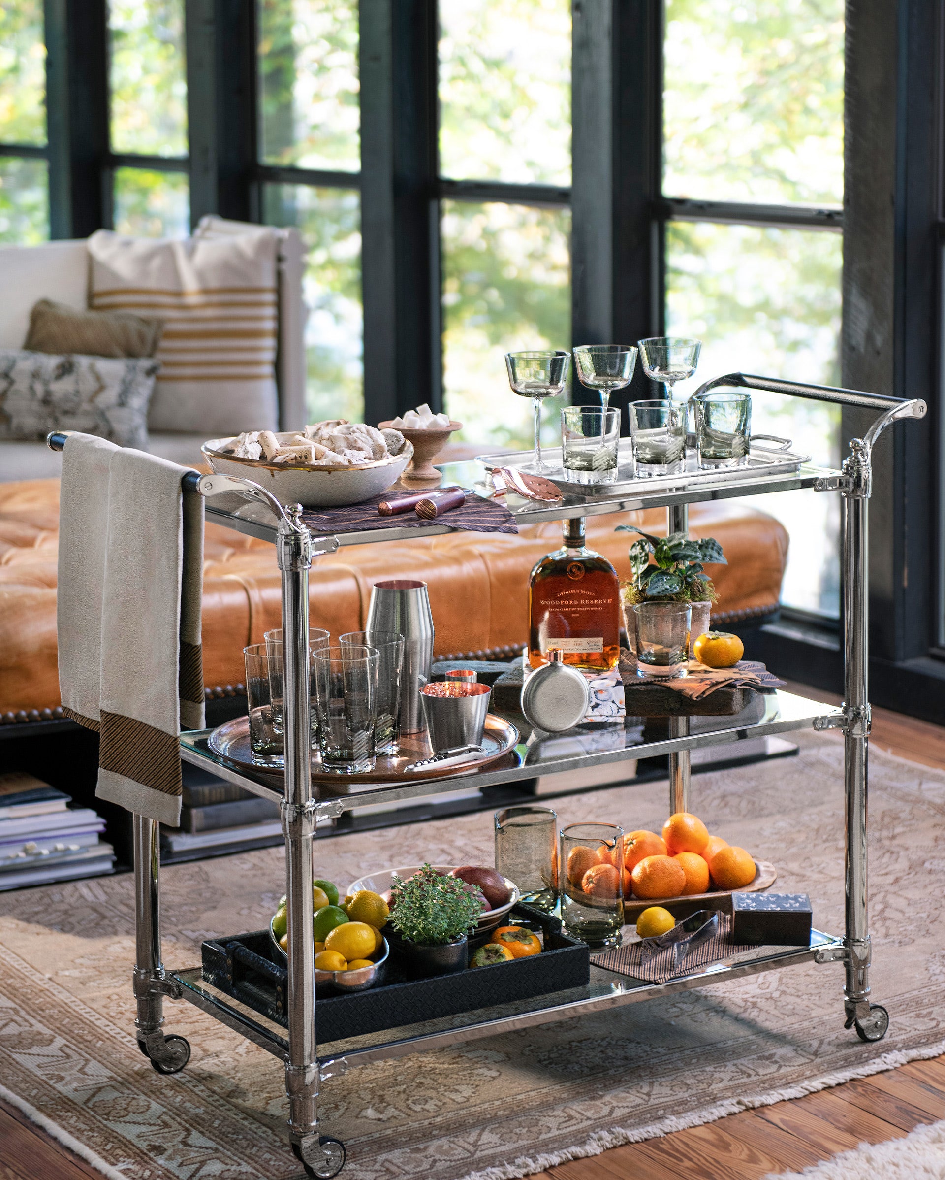 A metal bar cart with three shelves holds glassware, a bottle of whiskey, a cocktail shaker, snacks, and bowls of citrus fruits. It stands on a rug in a sunlit living room with large windows and a leather sofa.