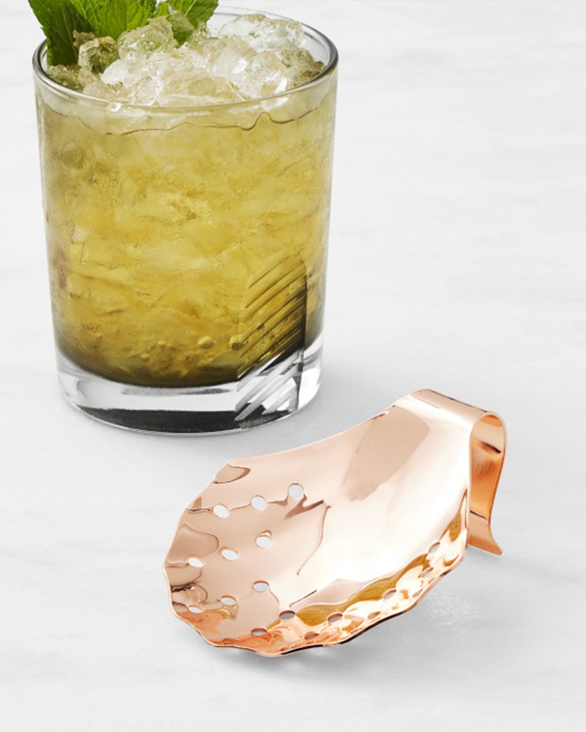 A glass of iced cocktail garnished with mint sits next to a hammered copper strainer with holes, resting on a white surface.