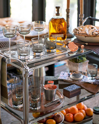 A silver bar cart with cocktail glasses, a bottle of whiskey, a bowl of cookies, a shaker, candles, a small potted plant, and a tray of fresh oranges on the lower shelf, set by a window.