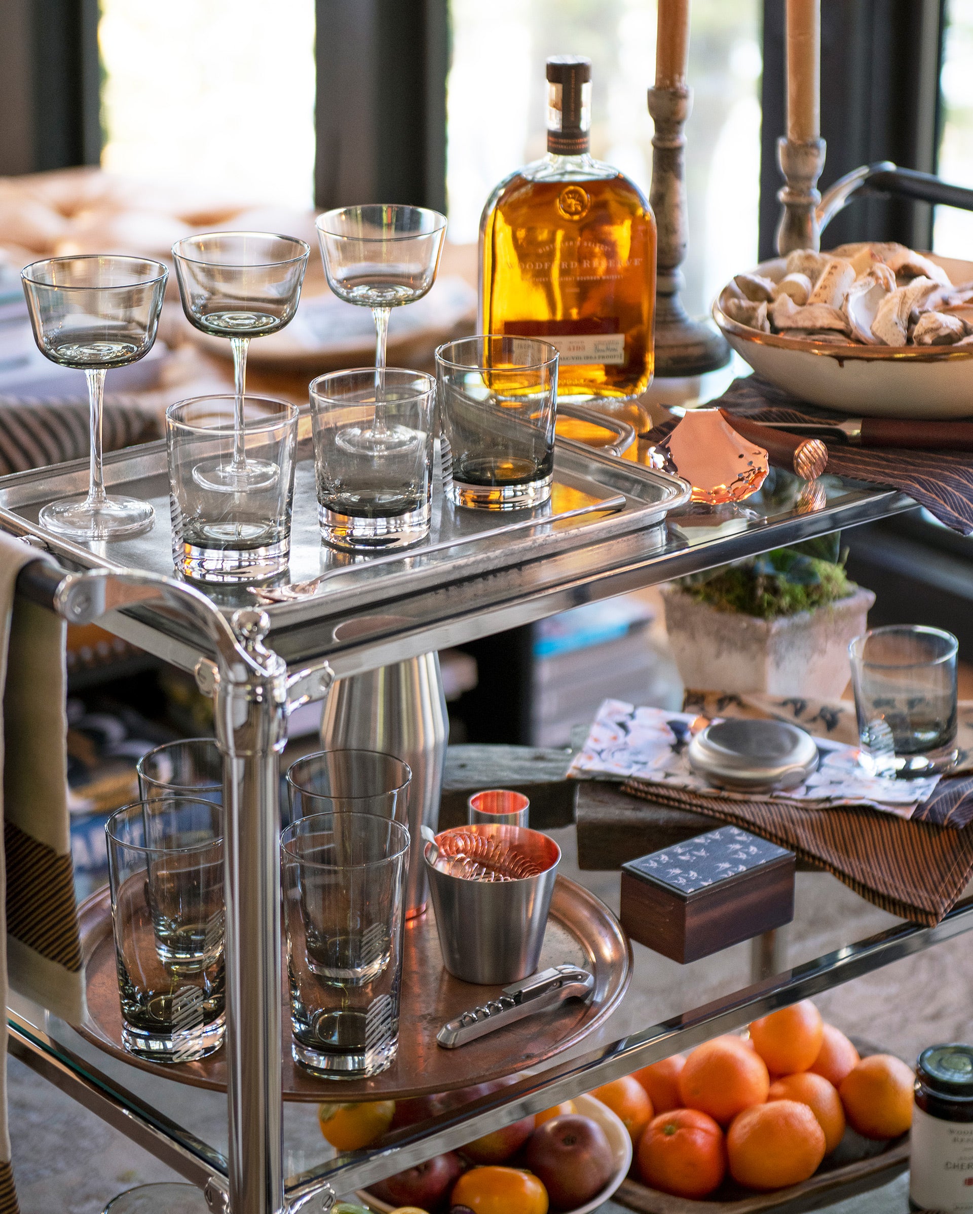 A bar cart holds various glasses, a bottle of liquor, a bowl of bread, a cocktail shaker, a copper cup, cocktail tools, and a small box, with a tray of oranges and other snacks on a table below.