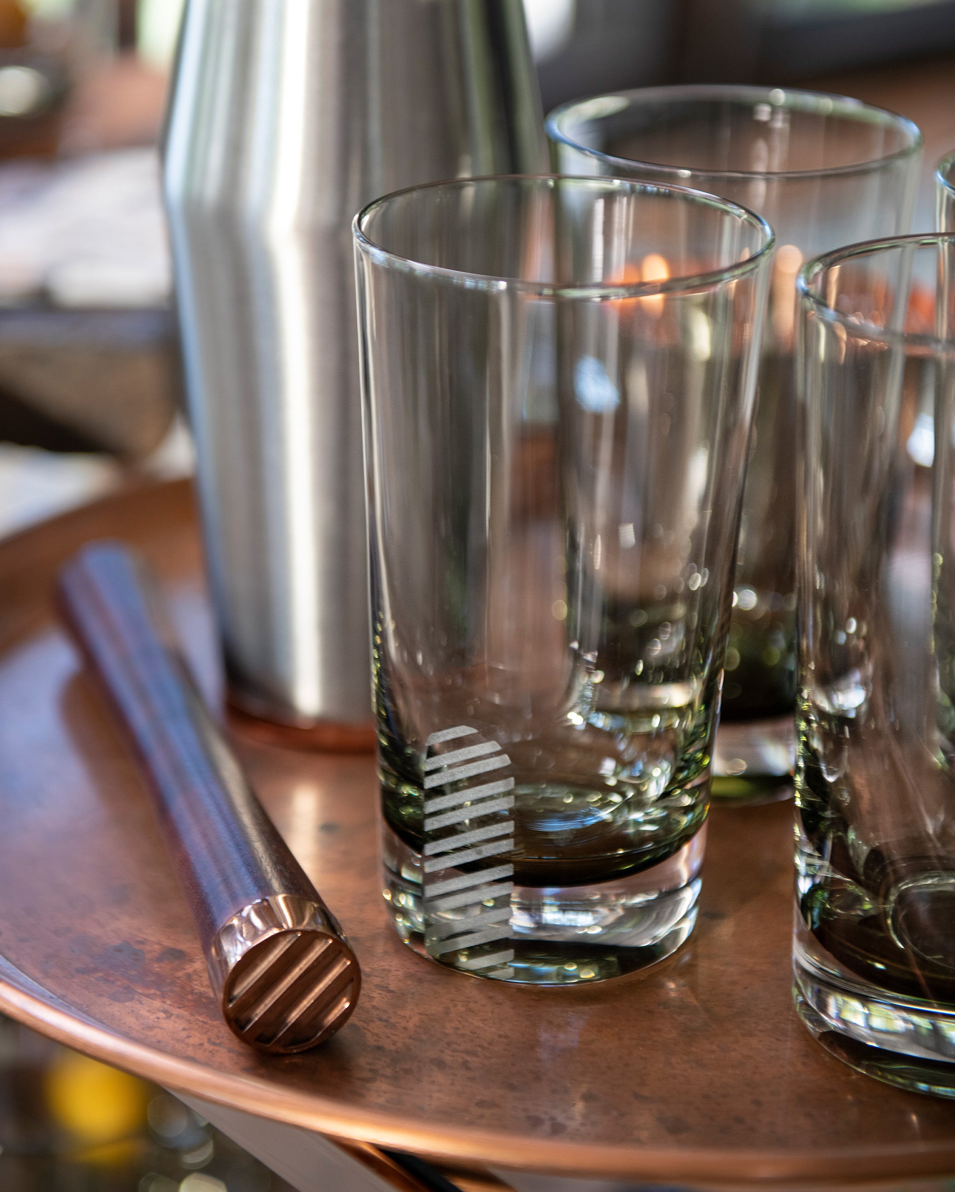 Clear glasses with a striped design sit on a copper tray next to a matching copper muddler and a stainless steel cocktail shaker, reflecting soft natural light.