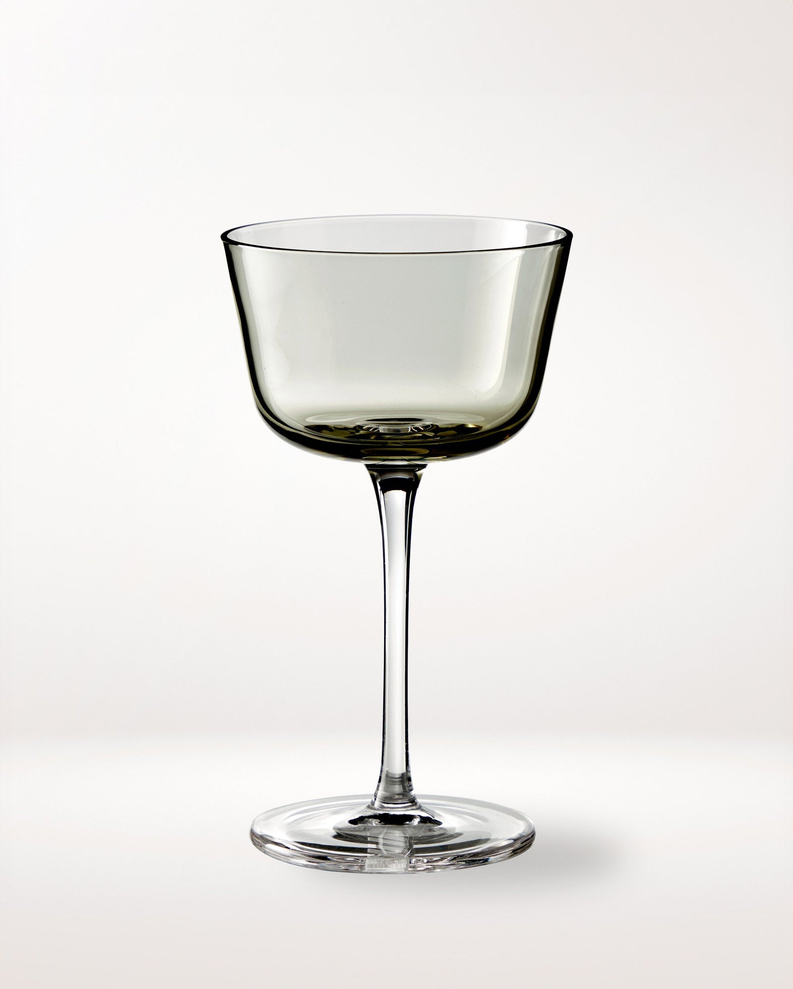 A single, elegant, clear wine glass with a wide bowl and long stem is centered on a plain, light background. The glass has a slightly smoky tint.