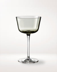 A single, elegant, clear wine glass with a wide bowl and long stem is centered on a plain, light background. The glass has a slightly smoky tint.