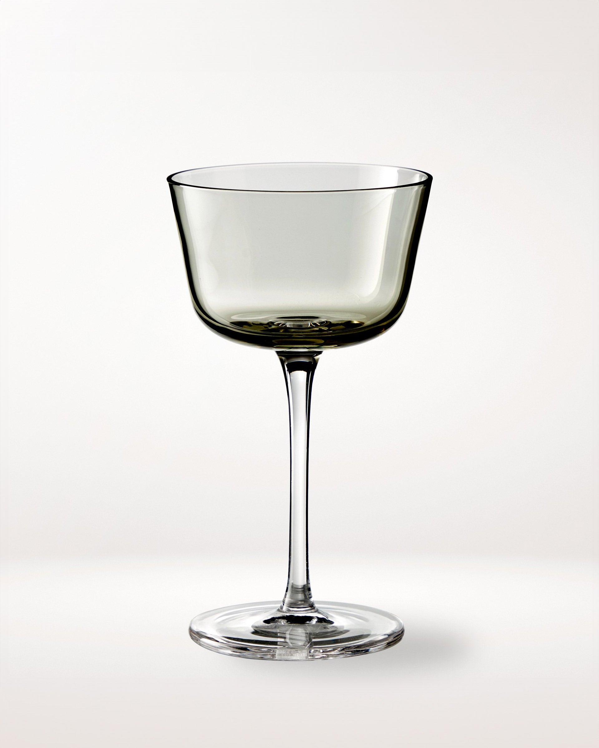 A single, elegant, clear wine glass with a wide bowl and long stem is centered on a plain, light background. The glass has a slightly smoky tint.